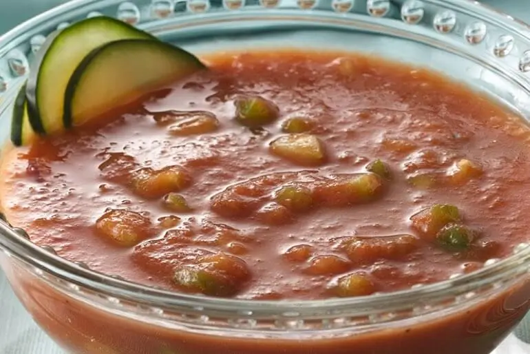Recette de soupe gazpacho avec des dés de tomates, de l'ail, du poivron vert et de l'oignon rouge, servie dans un bol en verre.