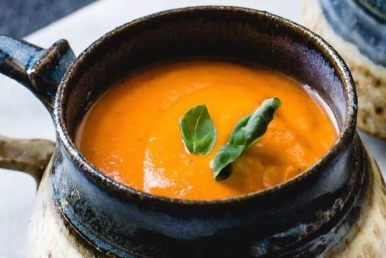 Recette de soupe crémeuse de tomates grillées à la noix de coco, garnie de basilic, dans un bol en céramique.