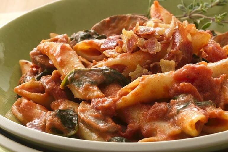 Penne pasta recipe with tomato sauce and mushrooms, topped with bacon, and garnished with a sprig of thyme.